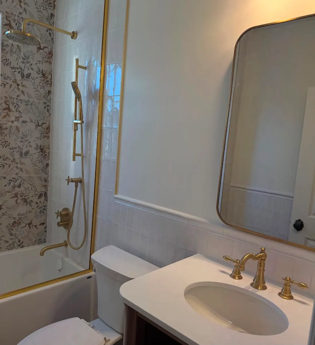 A finished bath with brushed gold shower fittings, floral wallpaper and a gold-framed mirror