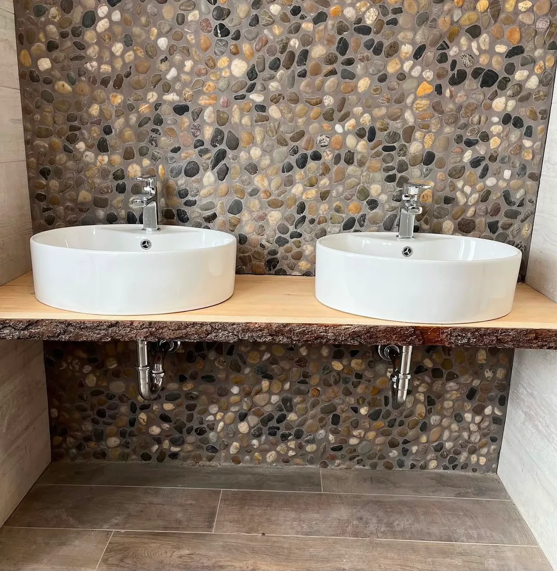 Twin white vessel basins on a live-edge timber shelf against a pebble mosaic wall